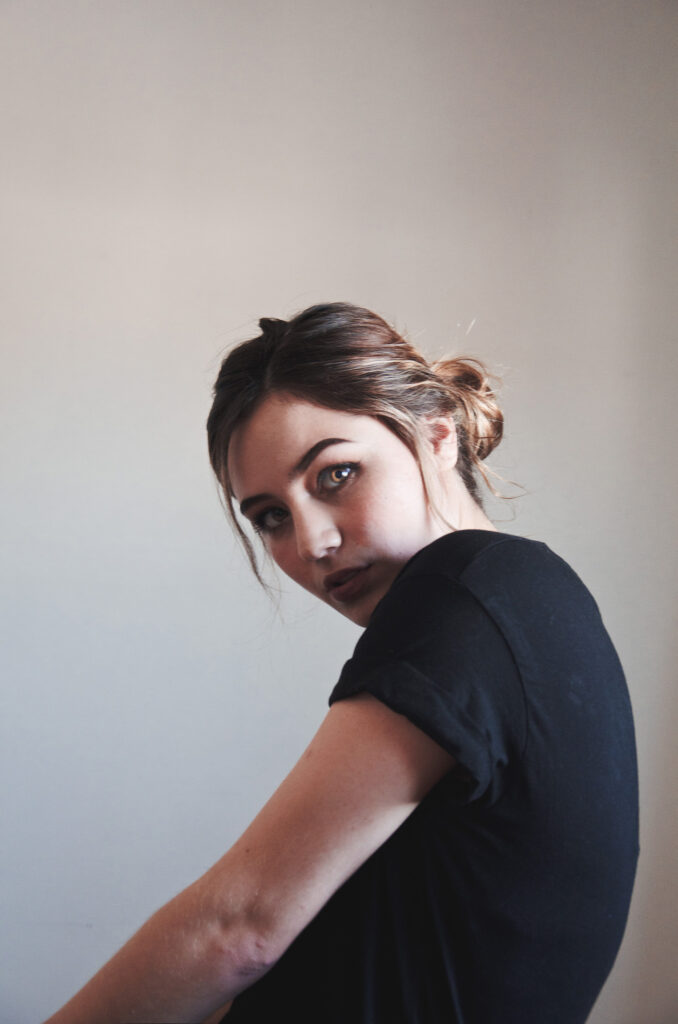 Darian Rojas a model with a striking gaze poses against a muted background wearing a black t-shirt with her hair tied back in a loose bun.